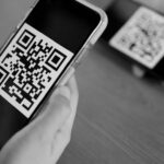 China's digital payment systems, enabling direct-to-consumer trading. Black and white photo of hands holding a personal device (iPhone) and scanning a QR code for payments.